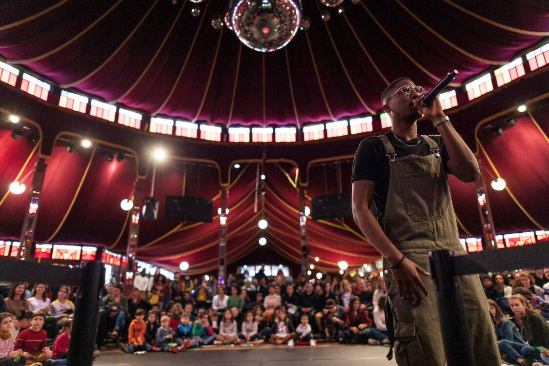 Personne tenant un micro en premier plan. Derrière une scène avec des enfants et un public par terre ou debout. Personne tenant un micro en premier plan. Derrière une scène avec des enfants et un public par terre ou debout.