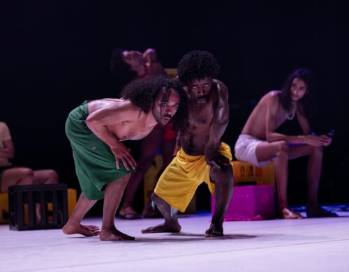 Deux danseurs en mouvement, un qui les regarde en fond.