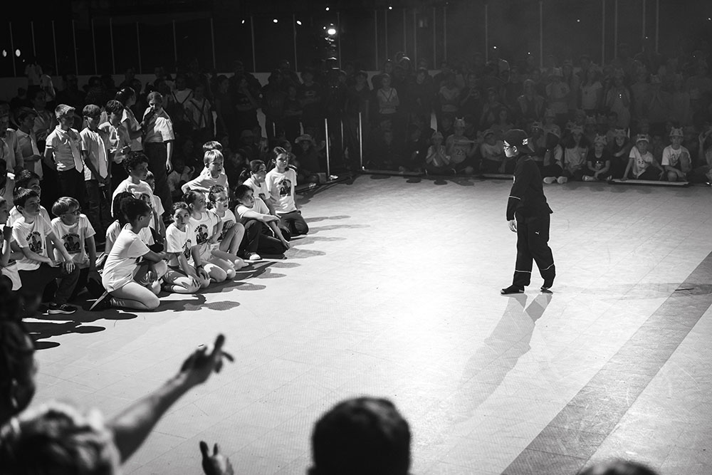 Photo en noir et blanc. Un enfant sur le devant face à d'autres qui danse durant une des battle. Photo en noir et blanc. Un enfant sur le devant face à d'autres qui danse durant une des battle.