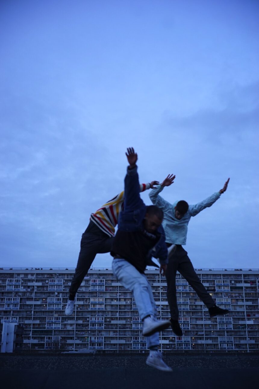 Trois danseurs en mouvement. Fond est un immeuble en large.