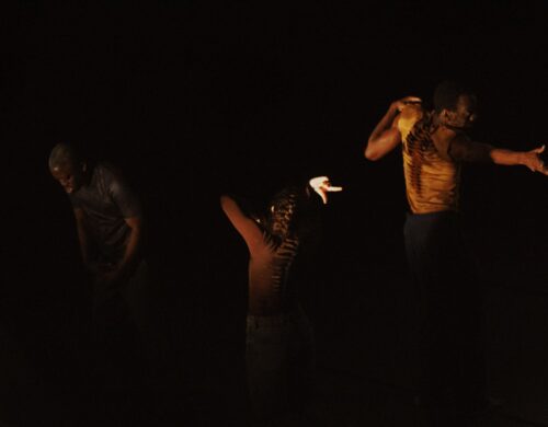 Trois danseurs sur scène en mouvement. Lumière jaune sur eux, fond noir.