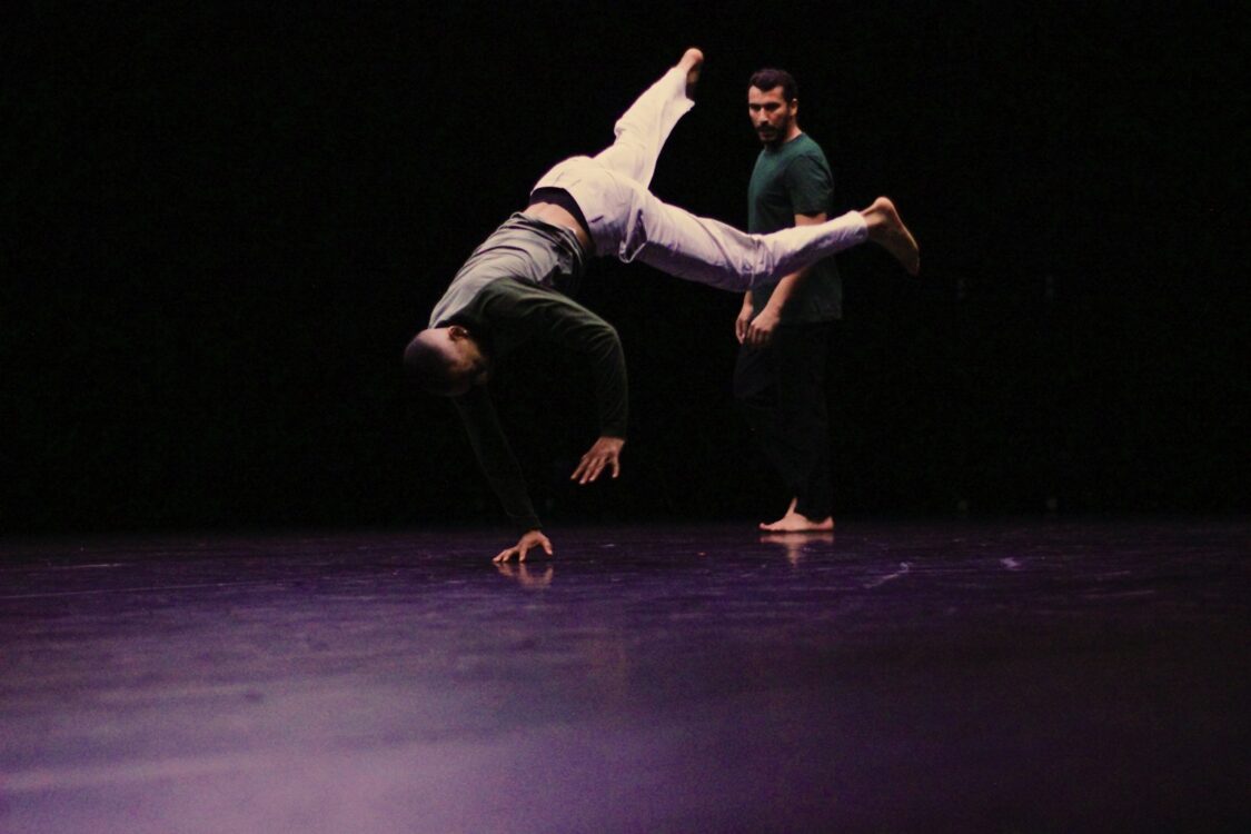 Un danseur en mouvement de breakdance, une main au sol, jambes en l'air. Un autre danseur dans le fond qui le regarde. Un danseur en mouvement de breakdance, une main au sol, jambes en l'air. Un autre danseur dans le fond qui le regarde.