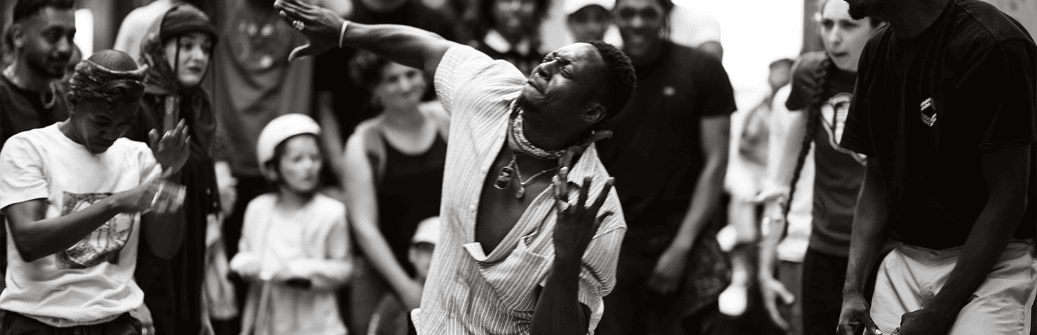 Photo en noir et blanc avec un danseur en premier plan et des personnes autour qui le regardent.