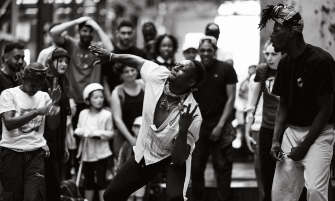 Photo en noir et blanc avec un danseur en premier plan et des personnes autour qui le regardent.