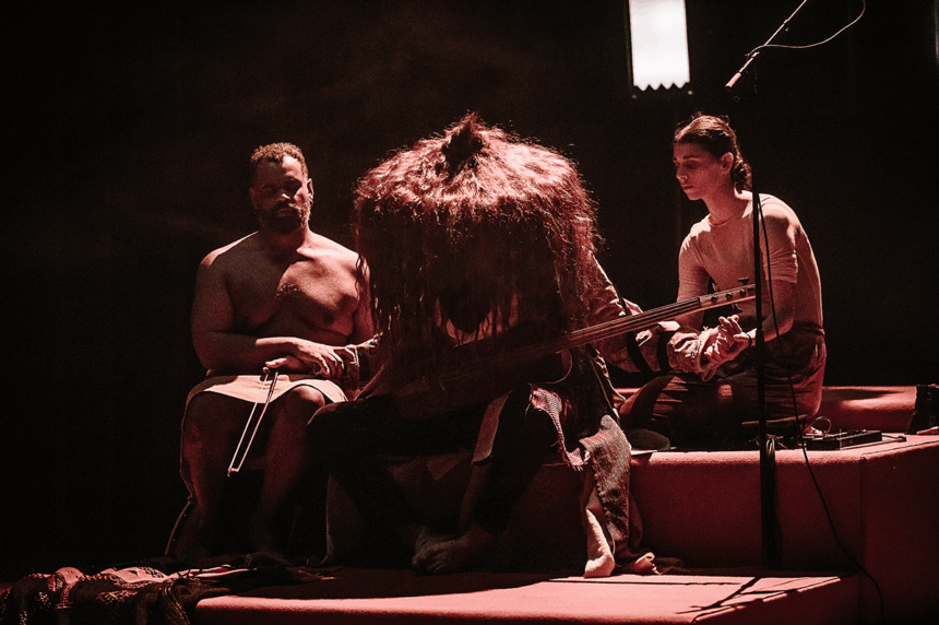 Trois personnes côte à côte. La personne de gauche a le regard perdu au loin. La personne au centre joue d'un instrument et porte un grand chapeau pointu fait de cheveux. La personne à droite regarde vers le bas.